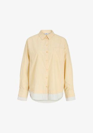 Lys beige langermet skjorte med knapper, spiss krage, brystlomme samt hvit nederkant og mansjetter på ensfarget bakgrunn.