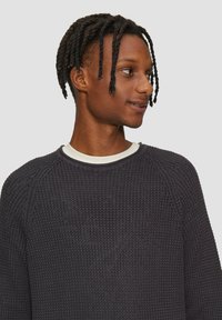 Dunkelgrauer gestrickter Pullover mit einem strukturierten Muster, geripptem Kragen und Raglanärmeln, der eine weiße Innenschicht am Ausschnitt sichtbar hat.