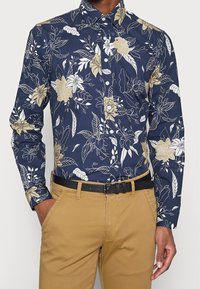 Camicia blu navy con bottoni e motivo floreale beige e oro. Presenta un colletto classico e maniche lunghe, abbinata a pantaloni kaki e cintura nera.