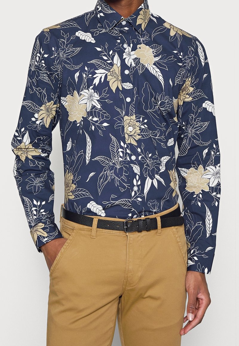 Marineblauw overhemd met knopen en een bloemenpatroon in crème en goud. Heeft een klassieke kraag en lange mouwen, gecombineerd met kaki broek en een zwarte riem.