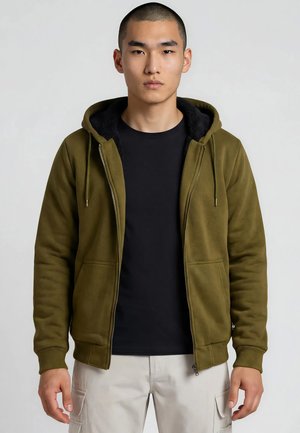 Uomo che indossa una giacca con cappuccio verde oliva sbottonata sopra una maglietta nera e pantaloni cargo chiari, in piedi davanti a uno sfondo semplice.