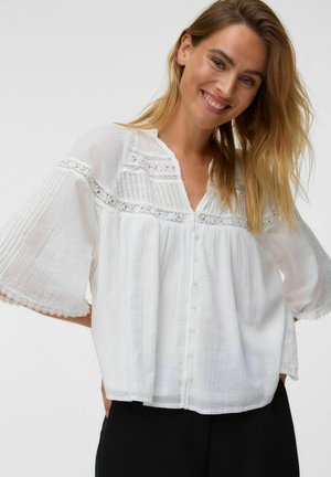 Femme souriante aux longs cheveux blonds porte un chemisier blanc ample avec des détails en dentelle et des boutons, associé à un pantalon noir sur un fond uni.