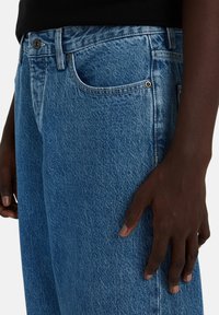 Dark-skinned person wearing blue denim jeans and a black top, with their right hand resting lightly on the thigh.