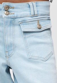 Ανοιχτού μπλε τζιν με ψηλή μέση, δύο μεταλλικά κουμπιά, τσέπη με καπάκι και κούμπωμα κουμπιού, και ορατές λεπτομέρειες ραφής.