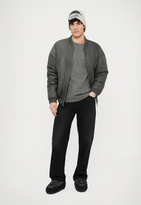 Pilka bombonešio striukė, pilka marškinėliai, juodos plačios kojų džinsai ir juodi raštuoti šlepetės. Jis dėvi šviesiai pilką kepuraitę. Paprasta balta fono spalva.