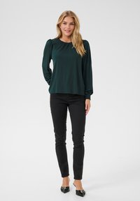 Blouse verte foncée à manches longues avec encolure froncée, tissu fluide et poignets ajustés, associée à un jean noir ajusté et des chaussures plates.