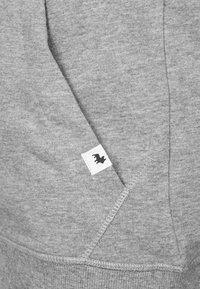 Felpa grigia con tasca frontale. Presenta una piccola etichetta nera con un logo. Il tessuto ha una texture liscia e una finitura leggermente spazzolata.