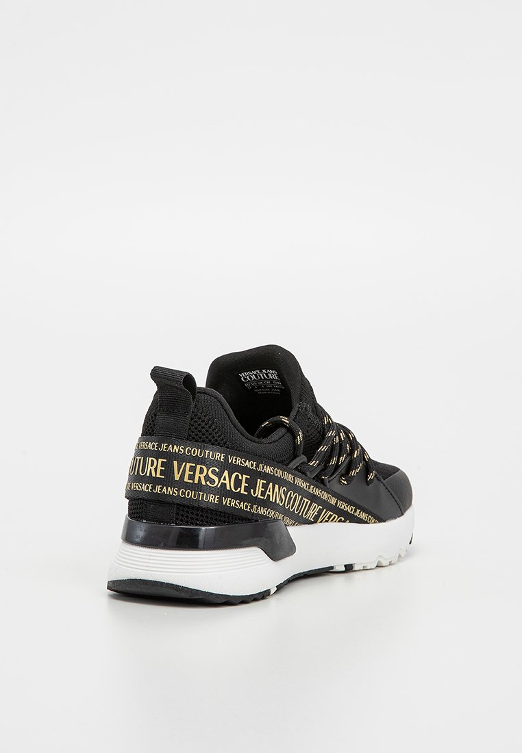 Schwarze und weiße Sneaker mit goldener "Versace Jeans Couture" Schriftzug auf dem Fersenriemen und schwarzen Schnürsenkeln mit goldenen Akzenten.