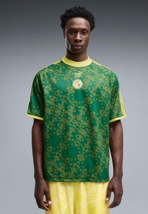 Bărbat purtând un tricou sport verde și galben cu model, cu logo Puma și un emblema pe piept, stând pe un fundal gri.