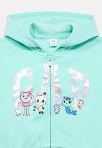 Mätovo zelená mikina na zips s reflexným logom "GAP" a kreslenými zvieracími postavičkami v rôznych farbách na prednej strane. Jemná tkanina.