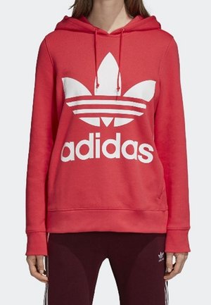 Personne portant un sweat à capuche rouge Adidas avec un grand logo Trefoil blanc et un pantalon Adidas rouge foncé assorti avec des bandes blanches.