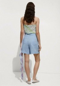 Svetle modré šortky s vysokým pasem a zadními kapsami, spárované s bezrukávovým topem s vzorem v zelené a bílé barvě. Modelka má dlouhé vlnité vlasy.