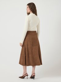 Mulher vestindo uma blusa de manga longa creme e uma saia midi de camurça castanha, de pé em sapatos de salto agulha castanhos, virada ligeiramente de lado diante de um fundo branco.