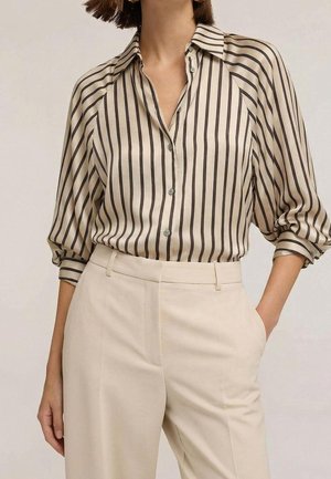 Kobieta w beżowych spodniach z wysokim stanem oraz kremowej bluzce z czarnymi pionowymi paskami i rękawami o długości do połowy przedramienia.