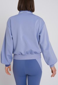 Maglione azzurro chiaro con fascia inferiore a coste, maniche ampie e logo discreto al colletto. Abbinato a leggings aderenti coordinati.