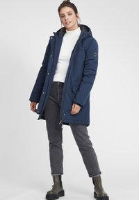 Mørkeblå polstret parka med hette, glidelås og trykknapper. Har en glatt stoffoverflate, sidelommer og et logomerke.