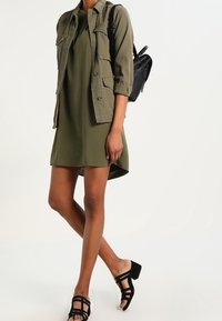 Robe verte olive avec un ourlet asymétrique, assortie à une veste utilitaire kaki dotée de plusieurs poches. Sandales à talons noires à lanières.