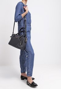 Svart texturerad handväska med dubbla handtag och ett avtagbart bälte. Modellen bär en denimjumpers och svarta loafers. Enligt vit bakgrund.