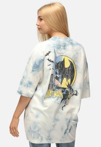Camiseta de algodón de gran tamaño en blanco con patrón de tie-dye azul, que cuenta con un gráfico de Batman en la parte trasera con detalles de murciélagos y el horizonte de la ciudad.