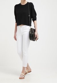 Suéter de punto negro recortado, jeans blancos ajustados, sandalias de tiras beige y un bolso cruzado negro con acentos metálicos. Diseño simple y moderno.