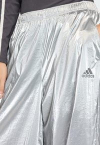 Ασημένια αθλητική φόρμα από γυαλιστερό, ελαφρύ υλικό με ελαστική μέση. Διαθέτει το μαύρο λογότυπο της Adidas στο πλάι.