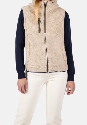 Persona que lleva un chaleco beige de forro polar con cremallera sobre una camiseta de manga larga azul marino y pantalones blancos, de pie frente a un fondo liso.