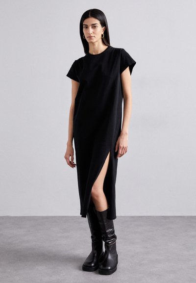 Schwarzes, kurzärmeliges Maxikleid mit einemSeitenschlitz, hergestellt aus weichem Stoff. Kombiniert mit hohen schwarzen Stiefeln, vor einem neutralen Hintergrund.