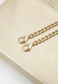 Zlatý kovový řetěz s dvěma svrchními sponami, připevněný na texturovaném zlatém podkladu, který zdůrazňuje hladký, lesklý povrch.