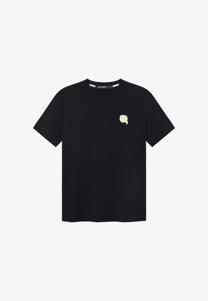 T-shirt noir à col en V et manches courtes, avec un petit motif abstrait beige en forme de visage sur le côté gauche de la poitrine et une étiquette à l'intérieur du col.