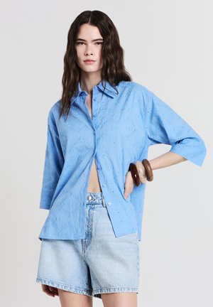 Frau in einem hellblauen, bestickten Hemd mit Knopfleiste und hellen Denim-Shorts, die mit der Hand auf der Hüfte posiert und Holzarmbänder am Handgelenk trägt.