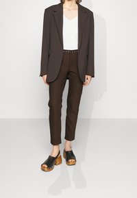 Blazer oversize marron, t-shirt blanc à col en V, pantalon marron cintré avec une ceinture à clous, et sabots noirs à bout ouvert avec des talons en bois.