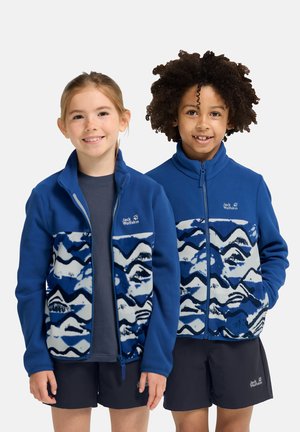 Twee lachende kinderen staan naast elkaar, beide dragen ze blauw gemêleerde jasjes met rits en zwarte shorts, tegen een effen achtergrond.