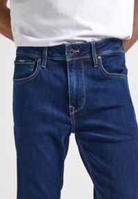 Temno modri denim kavbojki z klasičnim oblikovanjem petih žepov, kontrastnim šivom in blagovno znamko na pasu.
