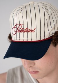 Baseballcap med kremfarget stoff, marineblå skygge og vertikale marineblå striper. "Forbudt" brodert i rødt foran.
