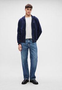 Hanorac bleumarin cu fermoar și șnururi, tricou alb cu guler rotund, blugi drepti de culoare albastru deschis și pantofi din piele negri.