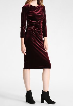 Vrouw met een nauwsluitende, knielange bordeauxrode fluwelen jurk met driekwartslagen en zwarte enkelhoge laarzen, staand tegen een witte achtergrond.
