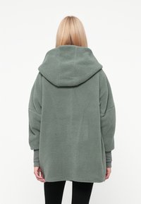 Weicher, übergroßer grüner Fleecepullover mit Kapuze, langen Ärmeln und einer strukturierten Oberfläche. Der untere Saum verläuft leicht rund.