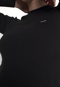 Primer plano de una persona que lleva un suéter negro de canalé con un pequeño logo blanco de Levi's en el pecho, con las manos descansando detrás del cuello.