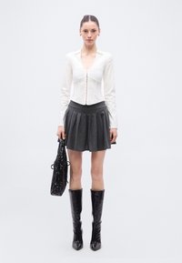 Bijela bluza s dugim rukavima i prednjim gumbima, uparena sa sivom plisiranom mini suknjom i crnim čizmama do koljena; model drži crnu torbu.