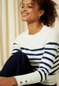 Maglione crema con strisce orizzontali navy, caratterizzato da un colletto rotondo e dettagli con bottoni sui polsini delle maniche. Indossato con jeans scuri.