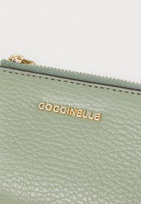 Carteira de pele texturizada verde-claro com o logótipo dourado "Coccinelle" e fecho de zip dourado. Apresenta uma borda em tecido contrastante.