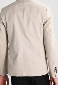 Blazer de un beige claro con una textura suave, corte entallado y una abertura simple en la parte posterior. Presenta cuatro botones negros en cada puño.