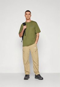 Λαδί βαμβακερό t-shirt, εκρού παντελόνι cargo με φερμουάρ στις τσέπες, μαύρα παπούτσια πεζοπορίας και μαύρο σακίδιο πλάτης. Απλή, λειτουργική εξωτερική ενδυμασία.