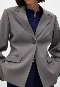 Pilkas švarkas su išdėstytais atlapais, vieno mygtuko uždarymu ir dviem priekinėmis kišenėmis. Dėvimas ant tamsiai mėlyno mygtukų marškinio. Lygus paviršius.