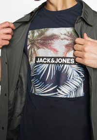 Muž v námořnickém tričku s designem palmy a logem "JACK & JONES", částečně zakrytý otevřenou šedou bundou.