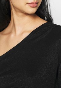Schwarzer, strukturierter Stoff mit leichtem Schimmer, mit einem diagonalen Ausschnitt und lockerer Passform, der den Bereich des Schlüsselbeins betont.