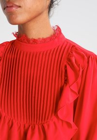 Blusa rossa con pieghe verticali, colletto arricciato e dettagli a volant sulle spalle. Texture liscia con una finitura vivace.