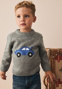 Grå stickad tröja med en blå bild av en bil på framsidan, ribbad halsringning och fåll, kombinerad med blå denimjeans. Brun resväska i bakgrunden.