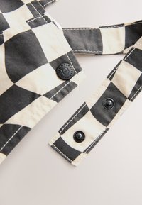 Tessuto a quadri nero e beige con una texture liscia. Presenta due bottoni neri e cuciture bianche a contrasto sui bordi.