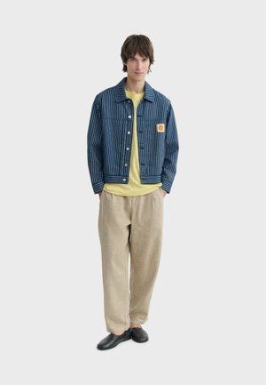 Jeune homme portant une veste rayée bleu et blanc, une chemise jaune, un pantalon beige et des chaussures noires sans lacets, debout devant un fond uni.